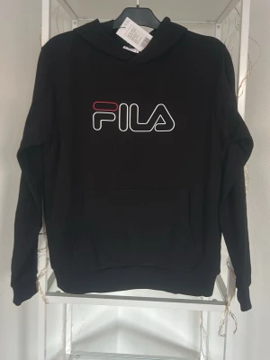 Svart hoodie från FILA med logga - Snygg svart hoodie från FILA med stor vit och röd logga på bröstet. Klassisk känguruficka framtill och huva. Perfekt för dig som gillar sportig och avslappnad stil. Passar dig som är 15-16 år och vill ha en enkel men trendig look.