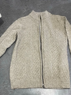 Beige stickad kofta med dragkedja - Säljer en beige stickad kofta med dragkedja framtill och snyggt fiskbensmönster. Koftan har ribbade muddar vid ärmslut och nederkant samt en högre krage. Perfekt för lager på lager och riktigt mysig.