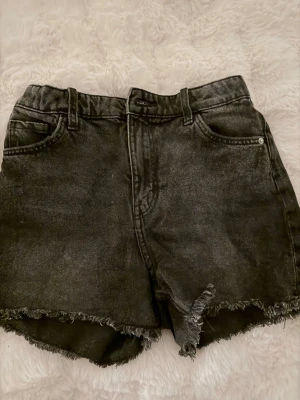 Svarta jeansshorts med rå kant - Säljer ett par svarta denimshorts med hög midja och fransig, rå kant nertill. Klassisk femficksmodell med dragkedja och knapp framtill. Perfekta till sommaren för en avslappnad och trendig look.