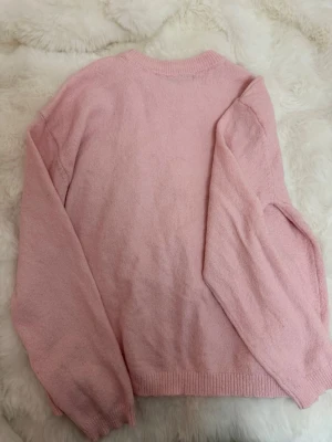 Ljusrosa stickad tröja - Mysig ljusrosa stickad tröja med rund halsringning och långa ärmar. Tröjan har ribbade muddar vid ärmslut och nederkant. Perfekt för att hålla värmen under kyliga dagar och enkel att matcha med olika outfits.
