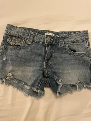 Blå jeansshorts med fransig kant - Säljer ett par klassiska blå jeansshorts med fransig kant och slitna detaljer. Shortsen har fem fickor, bälteshällor och knappgylf. Perfekta för varma dagar och en avslappnad stil.