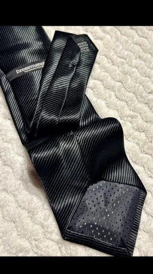 Svart slips från Dressmann - Snygg svart slips från Dressmann med diskret randigt mönster i satinliknande material. Slipsen har en klassisk form och känns stilren och enkel att matcha till olika skjortor och kavajer. Perfekt accessoar för att lyfta din outfit.
