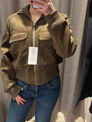 Olivgrön bomberjacka från Zara - Snygg olivgrön bomberjacka från ZARA i mockaimitation. Jackan har två stora bröstfickor med lock, ribbade muddar vid ärmslut och midja samt dragkedja framtill. Perfekt för dig som vill ha en trendig och stilren look. Jackan är slutsåld hos ZARA, och den är helt oanvänd med prislappen på.