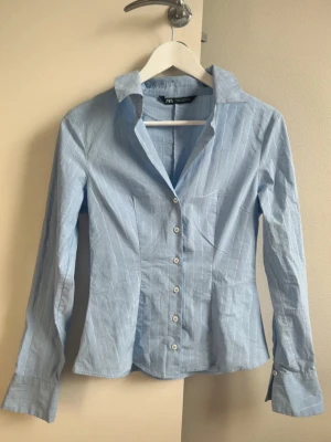 Ljusblå randig skjorta från Zara - Snygg ljusblå skjorta från Zara med vita tunna ränder och klassisk krage. Skjortan har lång ärm, figurnära passform och knäppning framtill med vita knappar. Perfekt till jeans eller kjol för en clean look. Den är i strl M, men som de flesta av Zaras kläder är den liten i strl💗 skulle säga att den passar xs/s