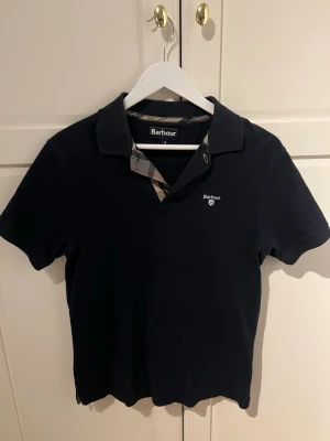 Marinblå pikétröja, Barbour - Säljer en marinblå pikétröja från Barbour med klassisk krage och korta ärmar. Tröjan har Barbour-logga broderad på bröstet och detaljer i rutigt tyg vid knäppningen. Storlek:M , Använd 2 gånger. Nypris 850kr