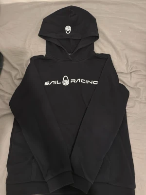 Sail racing Hoodie - Svart hoodie från Sail Racing med vit logga och text på bröstet samt logga på huvan. Klassisk känguruficka framtill och ribbade muddar vid ärmar och nederkant. Tillverkad i mjukt material som känns skönt mot huden. Perfekt för en avslappnad och sportig stil.