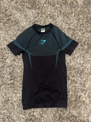 Gymshark onyx  - Snygg svart träningströja från Gymshark med korta ärmar och coola turkosa detaljer på axlar och bröst. Tillverkad i ett stretchigt funktionsmaterial som andas, perfekt för gymmet eller löpning. Gymshark-logga på bröstet och sportig design både fram och bak.