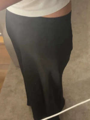 Svart långkjol i satin-look - Säljer en svart långkjol med snygg satin-finish. Kjolen har en rak och enkel passform som ger en clean och stilren vibe. Perfekt att matcha med crop tops eller stickade tröjor för en trendig look.