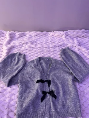 Grå kofta med svarta rosetter - Supersöt grå kofta med två svarta rosetter framtill. Koftan har v-ringning, långa ärmar och ribbade muddar. Perfekt för dig som gillar en gullig och trendig stil. Materialet känns mjukt och mysigt.