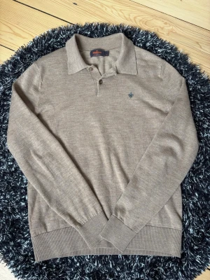 Beige pikétröja från Morris - Hej, säljer en klassisk beige, brun pikétröja från Morris med lång ärm och krage. Tröjan har två knappar vid halsen och en diskret blå lilje-logga broderad på bröstet. Tillverkad i mjukt, tunt stickat material som passar perfekt till en avslappnad men stilren look. Är i väldigt bra skick, hör av er vid frågor eller funderingar! 