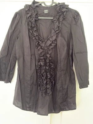 Svart volangblus från Esprit - Snygg svart blus från Esprit Collection med volangdetaljer längs ringningen och framtill. Blusen har trekvartsärm och är tillverkad i ett lätt tyg som ger ett elegant intryck. Perfekt för dig som gillar en chic och trendig stil.