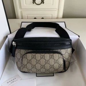 Beige axelväska med mönster - Snygg beige axelväska med klassiskt mönster i canvas och svarta detaljer i läder. Väskan har en justerbar axelrem, dragkedja upptill och en praktisk framficka med dragkedja. Perfekt för dig som gillar stilrena och lyxiga accessoarer.