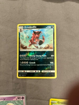 Pokémonkort Krookodile holografisk - Säljer ett holografiskt Pokémonkort med Krookodile. Kortet har en färgstark illustration av Krookodile i rött och svart mot en blå bakgrund. Text och detaljer är på engelska och ramen är gul. Perfekt för samlare eller Pokémonfans.