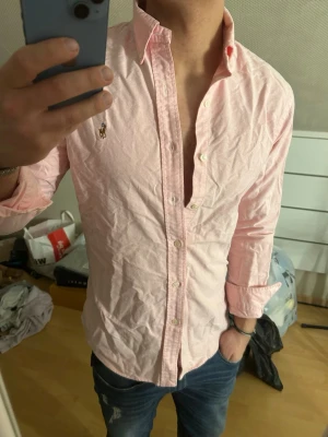 Ralph Lauren skjorta - Säljer en ljusrosa skjorta från Ralph Lauren med klassisk krage och knappar framtill. Skjortan har lång ärm och den ikoniska broderade loggan på bröstet. Tillverkad i bomull och har en normal passform. Perfekt för en stilren och avslappnad look.