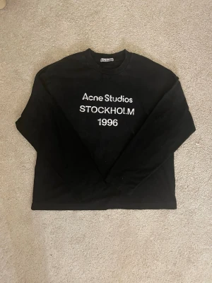 Svart långärmad topp Acne Studios - Svart långärmad topp från Acne Studios med vit text 'Acne Studios STOCKHOLM 1996' på bröstet. Klassisk rund halsringning och rak passform. Perfekt för dig som gillar stilrena och trendiga plagg med tydlig logga.