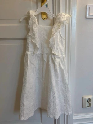 Vit klänning med volangdetaljer - Söt vit klänning med breda axelband och volangdetaljer framtill. Klänningen har en lätt utställd kjol och är tillverkad i ett strukturerat tyg som ger en elegant känsla. Perfekt för dig som gillar romantisk och feminin stil.