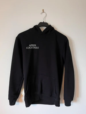 Svart hoodie Après Cocktails Club - Svart hoodie med vit text 'APRES COCKTAILS' på bröstet. Klassisk modell med huva och stor magficka. Tillverkad i mjukt material, perfekt för en avslappnad och trendig look. Passar dig som gillar stilrena och enkla plagg. Det är storlek xs men passar mer som S eller M