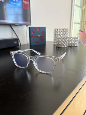 Transparenta solglasögon med silverdetaljer - Snygga solglasögon med transparent båge och fyrkantiga mörka linser. Silverfärgade detaljer med stjärnmönster på sidorna ger en unik touch. Perfekt för dig som vill sticka ut och ha en cool look i sommar.