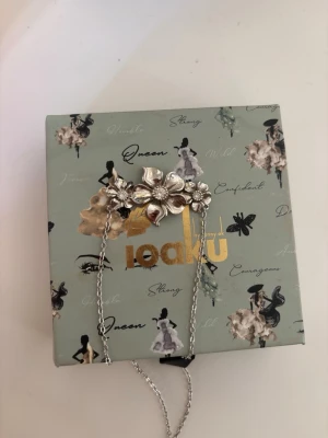 Blommigt halsband i silver från Ioaku - Säljer ett snyggt halsband från Ioaku med blommor i silverfärg. Halsbandet har en tunn kedja och ett stort blommotiv i mitten med flera detaljerade blomblad. Perfekt accessoar för att lyfta din outfit.