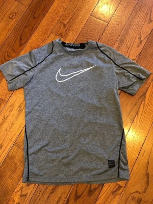 Grå Nike Pro t-shirt med logga - Sportig grå t-shirt från Nike Pro med svart kontrastsöm och stor vit Nike-logga på bröstet. Tillverkad i ett lätt och funktionellt material som passar perfekt för träning. Kortärmad och med rund halsringning.