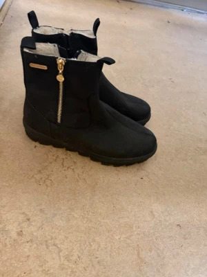 Svarta fodrade stövletter med dragkedja - Svarta stövletter med grov sula och guldfärgad dragkedja på sidan. Insidan är fodrad med ljust, mjukt material för extra värme. Snygg och enkel design med liten logodetalj på sidan. Perfekta för kyligare dagar.