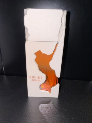 Ravine Ginger parfymflaska - Unik parfymflaska från Ravine med doften Ginger. Flaskan har en vit, kantig design med ett konstnärligt utskuret fönster som visar den orangefärgade vätskan inuti. Tillverkad i U.A.E. Perfekt för dig som gillar snygg och modern design.