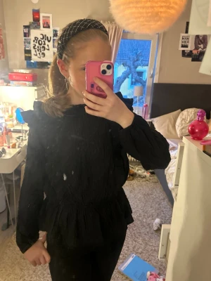 Svart volangblus med lång ärm - Trendig svart blus med volangdetaljer på axlar och midja. Blusen har lång ärm och smockad midja som ger en snygg siluett. Perfekt till både jeans och kjol för en cool look. Materialet ser ut att vara lätt och bekvämt.strl 158 men passar bra på mig som är xs-s