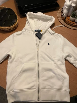 Vit hoodie från Polo Ralph Lauren - Snygg vit hoodie med st M från Polo Ralph Lauren med dragkedja framtill och klassisk blå logga på bröstet. .