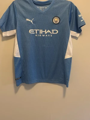 Manchester City matchtröja Grealish - Snygg ljusblå Manchester City matchtröja från Puma med Grealish och nummer 19 på ryggen. Tröjan har vita detaljer på sidorna, klubbmärke och sponsorlogga framtill samt Premier League-märke på ärmen. Perfekt för fotbollsfans!