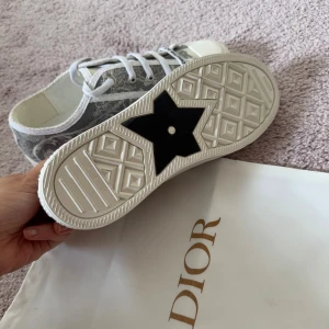 Gråvita sneakers från Dior - Snygga sneakers från Dior med gråvitt broderat mönster och vita snören. Skorna har en vit platt sula med svart stjärndetalj undertill och rund tå. Märkeslogga på sidan och tungan. Perfekta för dig som vill ha en exklusiv och trendig look. Passar de som har 39/40, har tyvärr inget kvitto kvar på de                                                               OBS KÖPAREN STÅR FÖR FRAKT!!! Skickar endast spårbart 