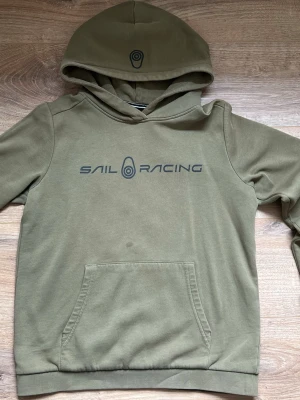Olivgrön hoodie från Sail Racing - Snygg olivgrön hoodie från Sail Racing med stor logga tryckt på bröstet och en mindre logga på huvan. Klassisk känguruficka framtill och mjukt material som känns skönt mot huden. Perfekt för en avslappnad och sportig stil.