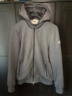 Grå hoodie från Moncler med dragkedja - Snygg grå hoodie från Moncler med hel dragkedja framtill, två fickor med dragkedja och klassisk Moncler-logga på ärmen. Hoodien har ribbade muddar och en stor huva. Perfekt för dig som vill ha en stilren och exklusiv look.