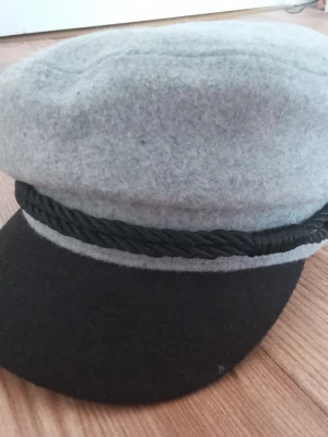 Grå och svart skepparmössa - Säljer en skepparmössa i grå filt med svart skärm och dekorativt svart snöre framtill. Mössan har en klassisk form och en knappdetalj på sidan. Köptes på Dollarstore. Observera att mössan är lite fläckig även om den är inte väl använd.