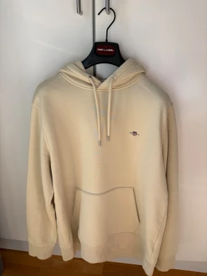GANT: Hoodie - Snygg beige hoodie från GANT med liten broderad logga på bröstet. Hoodien har huva med dragsko och en stor magficka framtill. Tillverkad i mjukt material som känns skönt mot huden. Perfekt för en avslappnad och stilren look. 🎉Hoodie köpt från GANT hemsidan för 1400kr och typ aldrig använd då för stor! 🎉För mer info skriv dm!