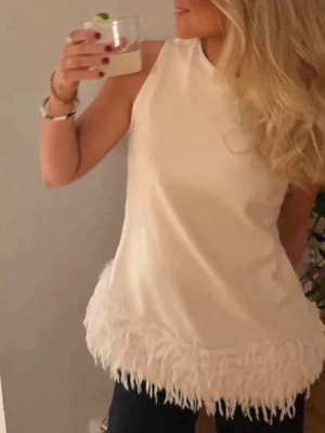 Vit ärmlös topp med fransar - Snygg vit ärmlös topp med rund hals och fransdetaljer längs nederkanten. Perfekt för dig som vill ha en enkel men ändå unik look. Toppen har en rak passform och är tillverkad i ett mjukt material som känns skönt mot huden.