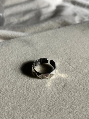 Silvrig justerbar ring med mönster - Säljer en stilren silvrig ring i metall med geometriskt mönster runt om. Ringen är öppen och justerbar i storlek, vilket gör den enkel att anpassa till olika fingrar. Perfekt för dig som gillar minimalistiska accessoarer med en twist.