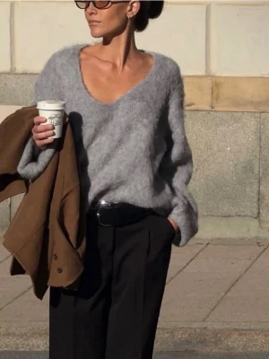 Ljusgrå fluffig stickad tröja - Mysig ljusgrå stickad tröja med djup v-ringning och lång ärm. Tröjan har en fluffig, mjuk struktur och avslappnad passform. Perfekt att styla med både jeans och kostymbyxor för en trendig look. 