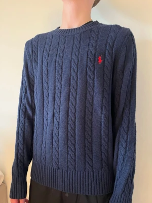 Mörkblå kabelstickad tröja Polo Ralph Lauren - Tvärfet mörkblå kabelstickad tröja från  Ralph Lauren med rund halsringning och röd broderad logga på bröstet. Tröjan är i jättebra skick och iprincip helt ny. Skickas snabbt och hör gärna av er vid minsta fundering!!!