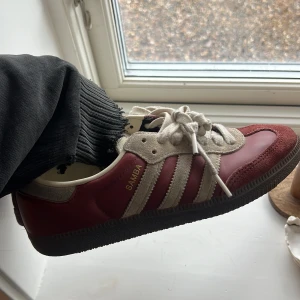 Adidas Samba vinröda sneakers - Adidas Samba sneakers i vinrött och beige mocka med klassiska vita ränder på sidan och gummisula. Köpte i höstas så nyskick förutom att de ljusa partierna blivit färjade av användning, kanske går att få bort men jag har inte testat, annasr prima! 