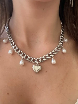 Silverfärgat halsband med pärlor och hjärta - Chunky halsband i silverfärgad metall med droppformade pärlor och ett hängande hjärta i mitten. Kedjan har en modern, bred länkdesign och pärlorna ger en söt kontrast till det tuffa metallutseendet. Perfekt för att lyfta vilken outfit som helst.