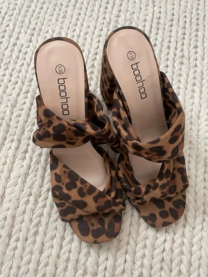 Leopardmönstrade sandaler från Boohoo - Säljer ett par trendiga sandaler från Boohoo med leopardmönster i brunt och svart. Skorna har bred klack och öppen tå, samt twistad rem över foten. Materialet är mjukt tyg med mockakänsla. Perfekta för dig som vill sticka ut med din stil.