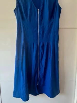 Blå ärmlös klänning med dragkedja - Snygg blå ärmlös klänning med v-ringning och dragkedja framtill. Klänningen har en lätt utställd passform och resår i ryggen för extra komfort. Perfekt för dig som vill ha en enkel men stilren look.