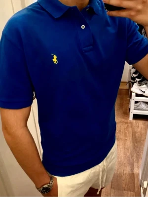 Mörkblå Piké från Ralph Lauren (Slim Fit) - Snygg blå pikétröja från Polo Ralph Lauren med gul broderad logga på bröstet. Klassisk krage, två knappar vid halsen och korta ärmar. Tillverkad i mjuk bomull och har en normal passform som sitter riktigt bra. Storlek Small slim fit. Snabb Leverans!