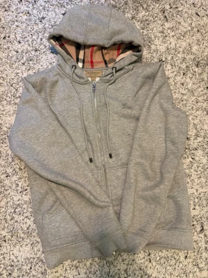 Grå hoodie från Burberry med dragkedja - Snygg grå hoodie från Burberry med klassiskt rutigt foder i huvan. Tröjan har dragkedja framtill, dragsko i huvan och ribbade muddar. Perfekt för en stilren och avslappnad look. Materialet är mjukt och bekvämt, troligen bomullsblandning. Priset kan diskuteras.