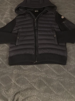 Svart dunväst med huva från Moncler - Snygg svart dunväst från Moncler med huva och stickade detaljer på ärmar och nederkant. Västen har dragkedja framtill, två fickor med tryckknapp och Moncler-logga på ärmen. Perfekt för lager-på-lager och streetstyle.