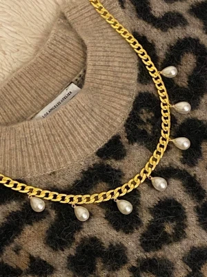 Guldhalsband med droppformade pärlor - Snyggt halsband i guldfärgad rostfritt stål. Kedjan är chunky och pryds av sju droppformade vita pärlor som hänger längs hela halsbandet. Perfekt för att ge din outfit en trendig och elegant touch.