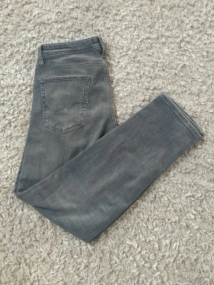 Grå jeans från Jack & Janes  - Säljer ett par snygga grå jeans från Jack & Jones med klassisk femficksmodell och diskret tvättad look. Jeansen har raka ben och silverfärgade nitar. Perfekta för dig som gillar stilrena och tidlösa plagg i garderoben.