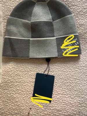 Gråmelerad rutmönstrad mössa - Snygg gråmelerad mössa med rutmönster i olika nyanser av grått. Mössan har vikt kant och diskret logga på sidan. Tillverkad i mjukt stickat material som håller dig varm under kalla dagar. Perfekt accessoar för en stilren och modern look.