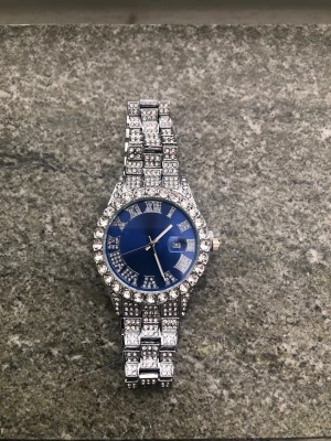 Klocka - Snygg klocka med glittrande stenar på både armband och boett. Urtavlan är blå med romerska siffror och datumvisning. Klockan har en lyxig känsla och passar perfekt för dig som gillar att sticka ut.