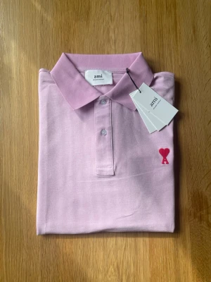 Rosa pikétröja från Ami Paris - Säljer en rosa pikétröja från Ami Paris med klassisk krage och knappar framtill. Tröjan har Ami:s ikoniska röda hjärtdetalj broderad på bröstet. Tillverkad i bomull för en skön och luftig känsla. Perfekt för en clean och stilren look.